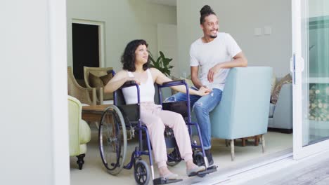 Happy-biracial-woman-in-wheelchair-and-smiling-male-partner-looking-out-of-terrace-in-living-room