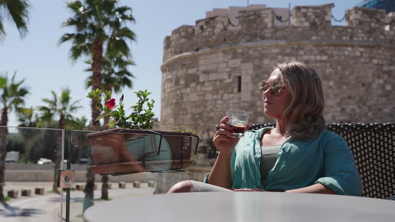 Woman with sunglasses and blue shirt drinks from glass near medieval stone castle with palm trees in background, Durres, Albania