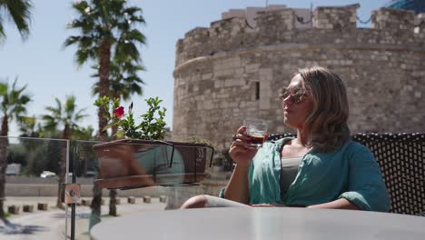 woman with sunglasses and blue shirt drinks from glass near medieval stone castle with palm trees in background, durres, albania