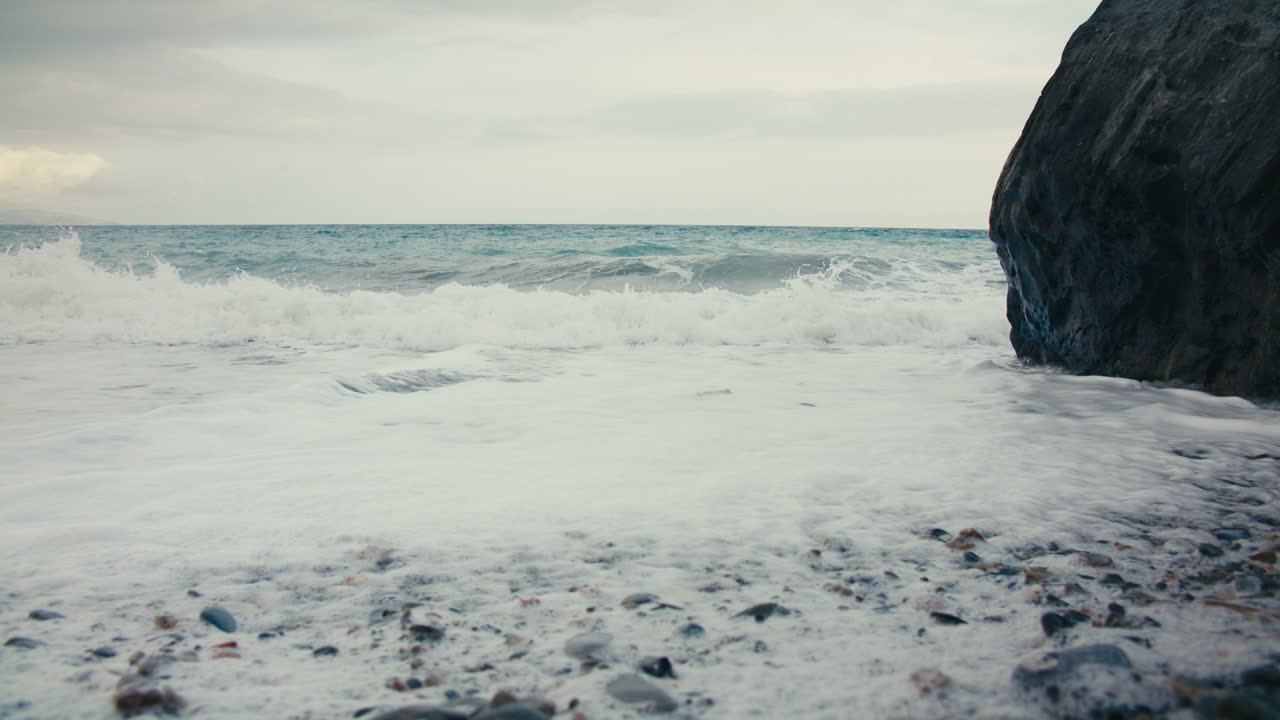 Low-angle shot of ocean wave approaching a rocky shore, with thick white foam covering the pebbled beach. A large dark cliff is visible on the right side. Overcast sky and horizon in the distance