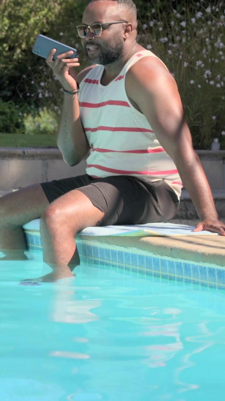Vertical video: Alternating man dipping feet and speaking into phone at poolside in striped tank