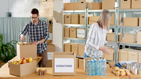 young caucasian male volunteer putting food in a donation box in charity warehouse while senior woman typing on laptop