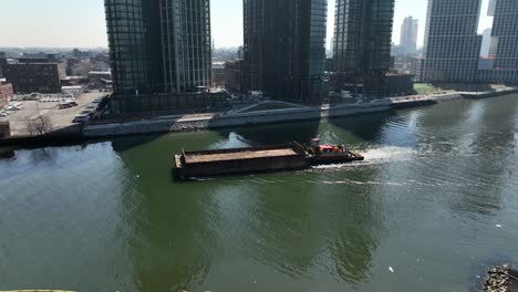 An-aerial-view-of-a-barge-sailing-down-Newtown-Creek-with-new-high-rise-apartment-buildings-in-Brooklyn,-NY-in-the-background-on-a-sunny-day