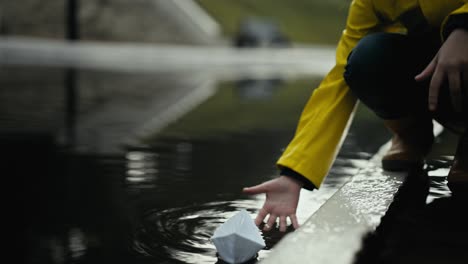 close-up una adolescente en una chaqueta amarilla lanza un barco de papel blanco en el agua a través de un gran charco durante la lluvia en la calle