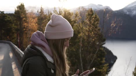 Frau-Mit-Beigem-Hut-Blickt-Von-Der-Straße-Aus-Auf-Den-Horizont-Und-Fotografiert-Mit-Ihrem-Smartphone-Die-Unglaublich-Schöne-Landschaft-Norwegens-Mit-Bergen,-Schneebedeckten-Gipfeln-Und-Einem-See.-Seitenansicht