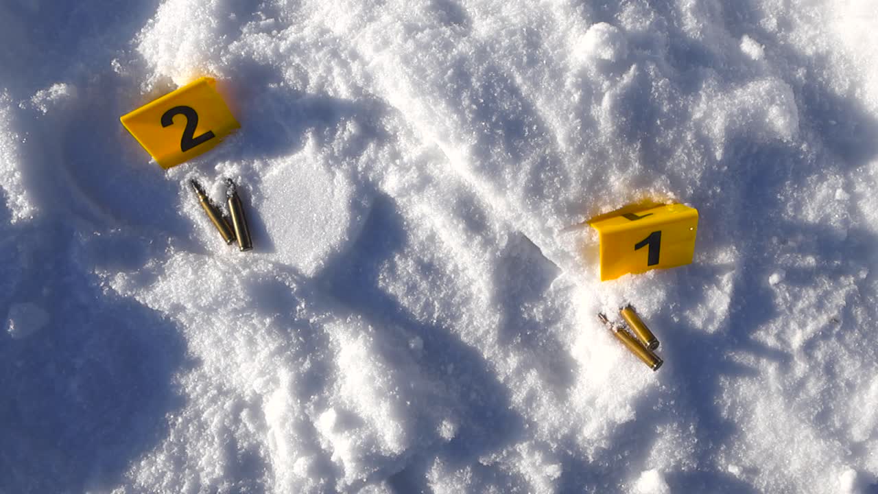 Una ripresa aerea si avvicina a marcatori numerati, bossoli in ottone e un'impronta visibile impressa nella neve brillante.