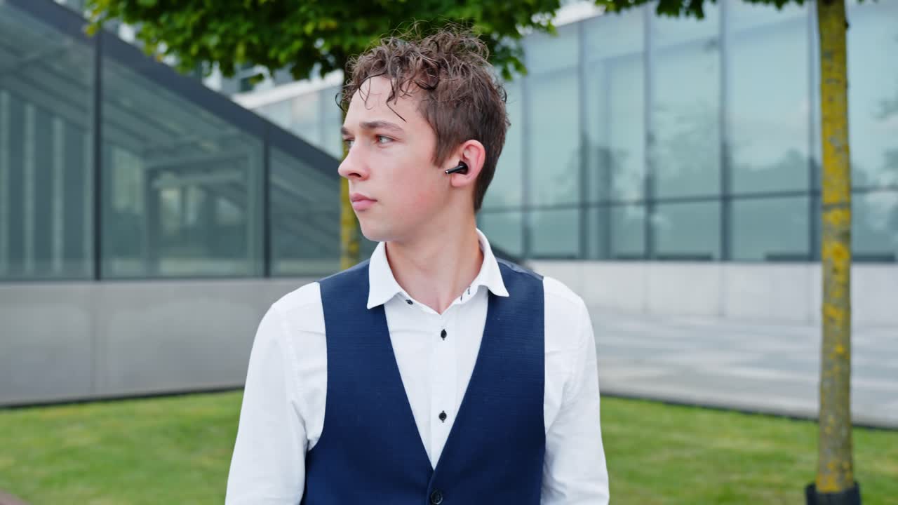 Young european man with an earbud in his ear, engaged in a call or listening to music or a podcast, feeling thoughtful and looking away outdoors in a modern urban environment, slow motion follow shot