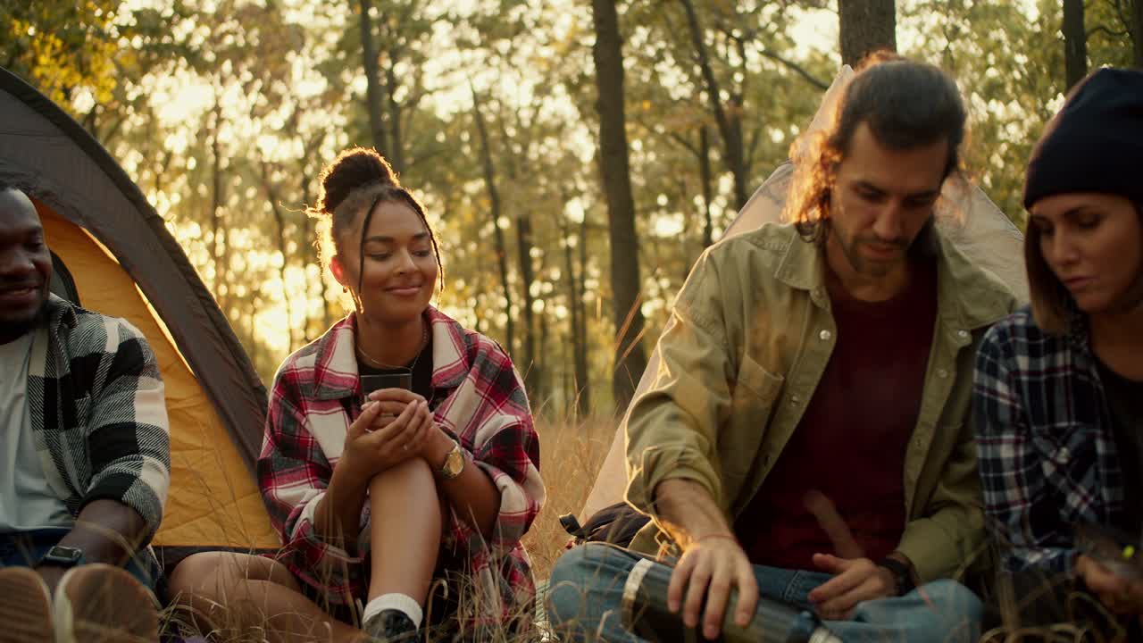 Close-up shot of a happy group of people on a hike in plaid shirts and hiking clothes pouring tea into glasses against the backdrop of a sunny summer forest. Using a thermos during a halt against the backdrop of tents