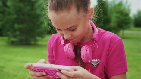 a joyful young girl in a pink shirt with stylish headphones smiles as she interacts with her smartphone in a vibrant outdoor setting filled with greenery