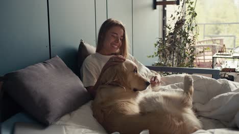 Happy-blonde-girl-petting-her-dog-in-bed-at-home-against-the-background-of-a-turquoise-shade.-Harmony,-happiness-and-joy