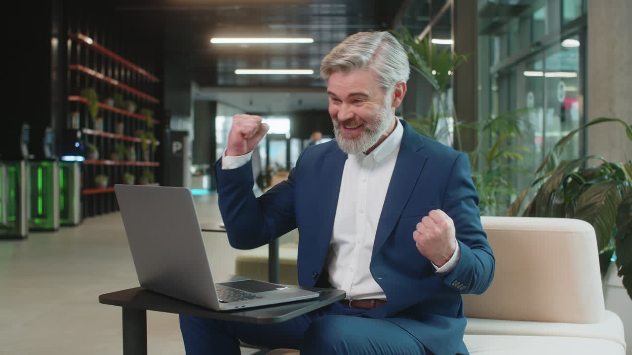 Caucasian bearded businessman working on laptop surprised by sudden victory in modern hotel lobby