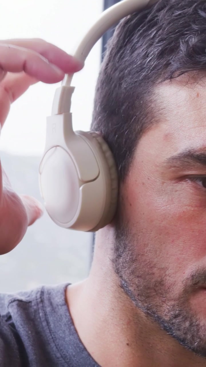 Video verticale: Uomo in maglietta grigia che controlla la cuffia, mettendosi delle cuffie beige in testa vicino alla finestra per ascoltare.