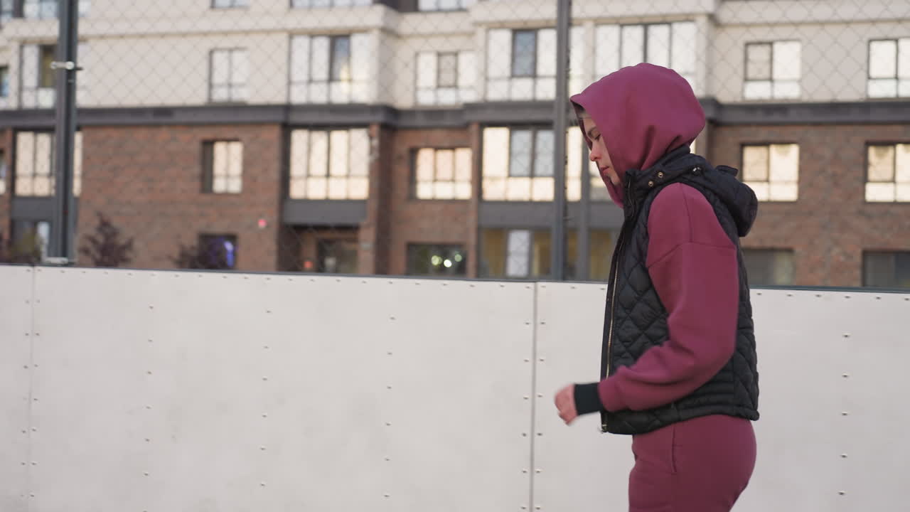 Side view of urban dweller in hoodie jogging around curved outdoor barbed wire court under pastel dusk sky, framed by chain link fence against apartment building backdrop glowing at sunset