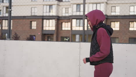side view of urban dweller in hoodie jogging around curved outdoor barbed wire court under pastel dusk sky, framed by chain link fence against apartment building backdrop glowing at sunset