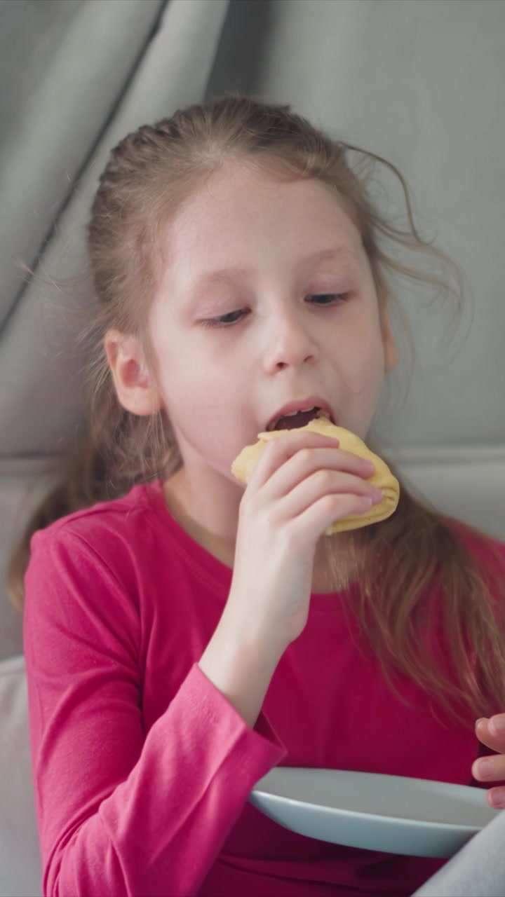 Close up of young girl eating sandwich while holding it in her hand. Girl wears pink shirt and sits at table with white plate visible. Soft lighting creates natural indoor setting