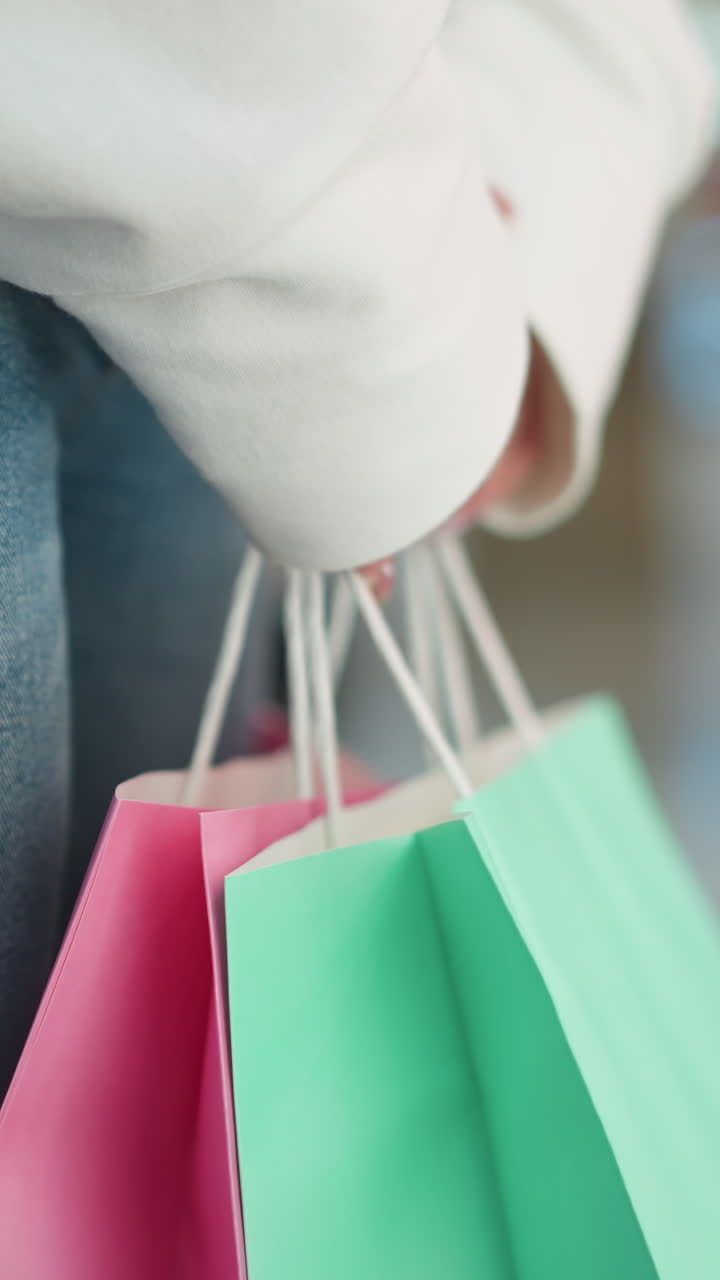 primer plano de una mujer con sudadera blanca y vaqueros de pie en el interior sosteniendo bolsas de compras de menta y rosa, fondo borroso con luces de colores y entorno comercial