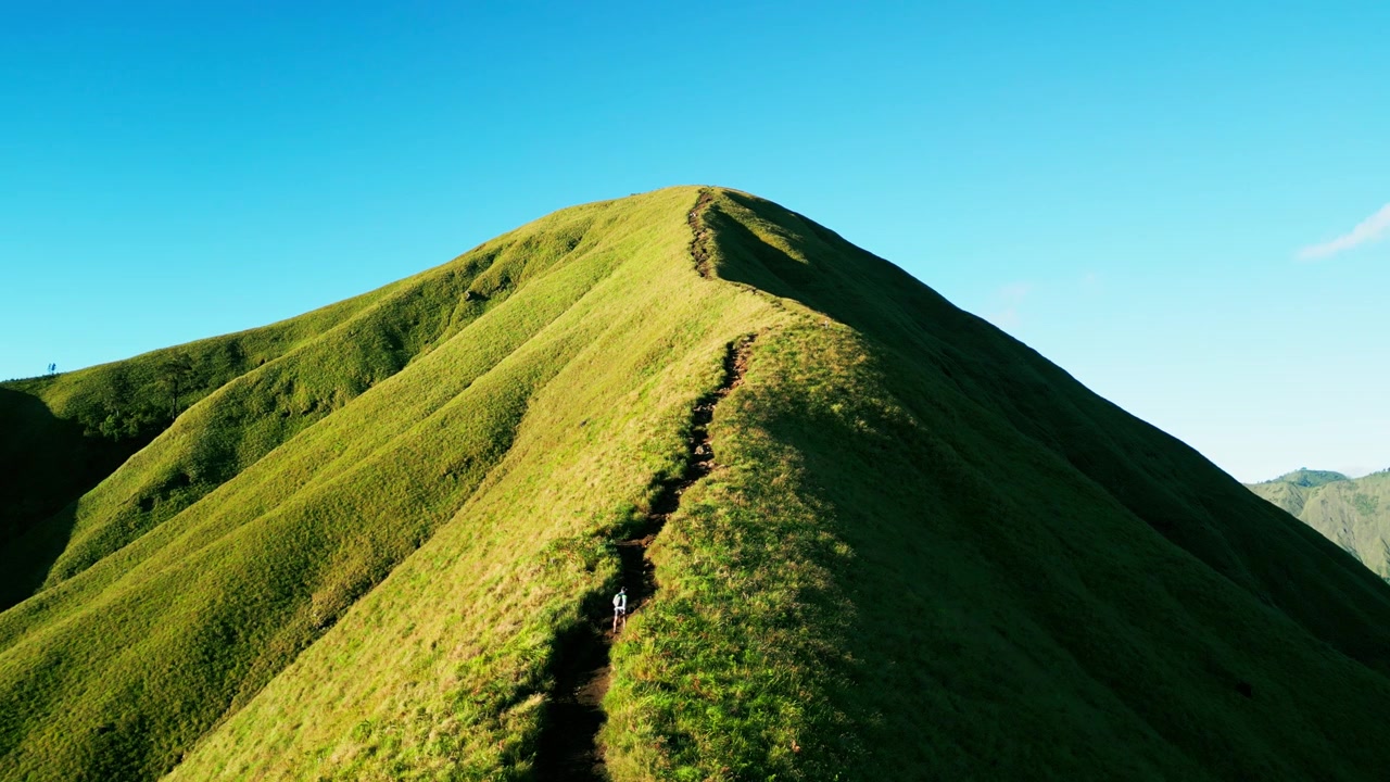 Avventuriero solitario sale su un ripido crinale montuoso erboso circondato da dolci colline verdi illuminate dal sole sotto un cielo azzurro limpido e soleggiato a Lombok, filmato aereo statico
