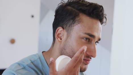 Happy-biracial-man-applying-face-cream-looking-in-bathroom-mirror,-slow-motion