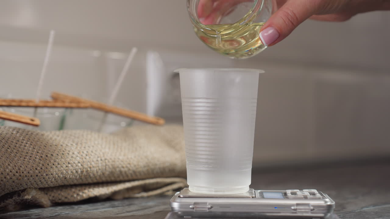 Close-up hand pouring baking liquid from glass cup into measuring cup on digital scale with soft cotton cloth and wooden wicks visible in background kitchen setting