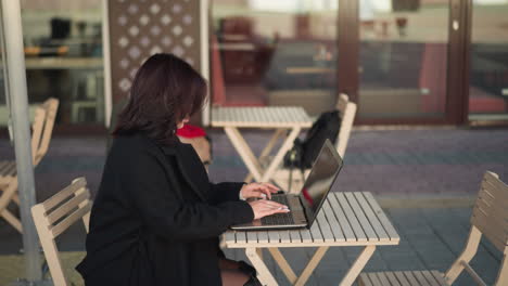 side view of woman typing on laptop outdoors in urban restaurant setting, wearing black coat, with glass reflecting restaurant interior and wooden chairs arranged neatly in modern environment