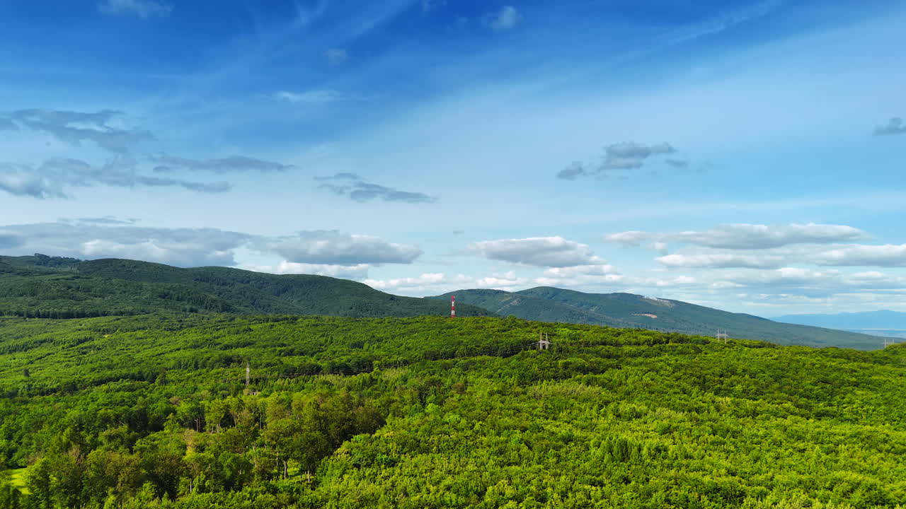 Lush green landscape under a bright sky. Vast greenery stretches out beneath a clear blue sky, with rolling hills in the background and scattered clouds above