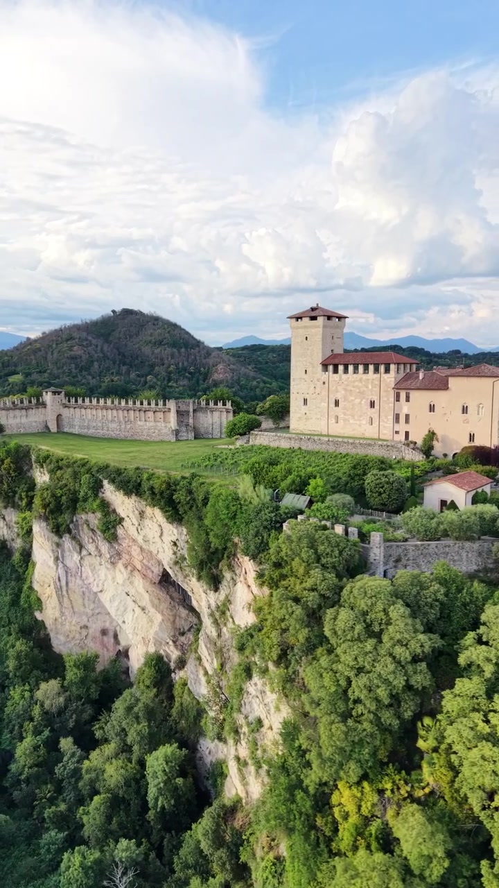 Il Castello Borromeo di Angera si erge sopra alberi lussureggianti sul bordo della scogliera con il Lago Maggiore, orbita aerea verticale di inquadratura.