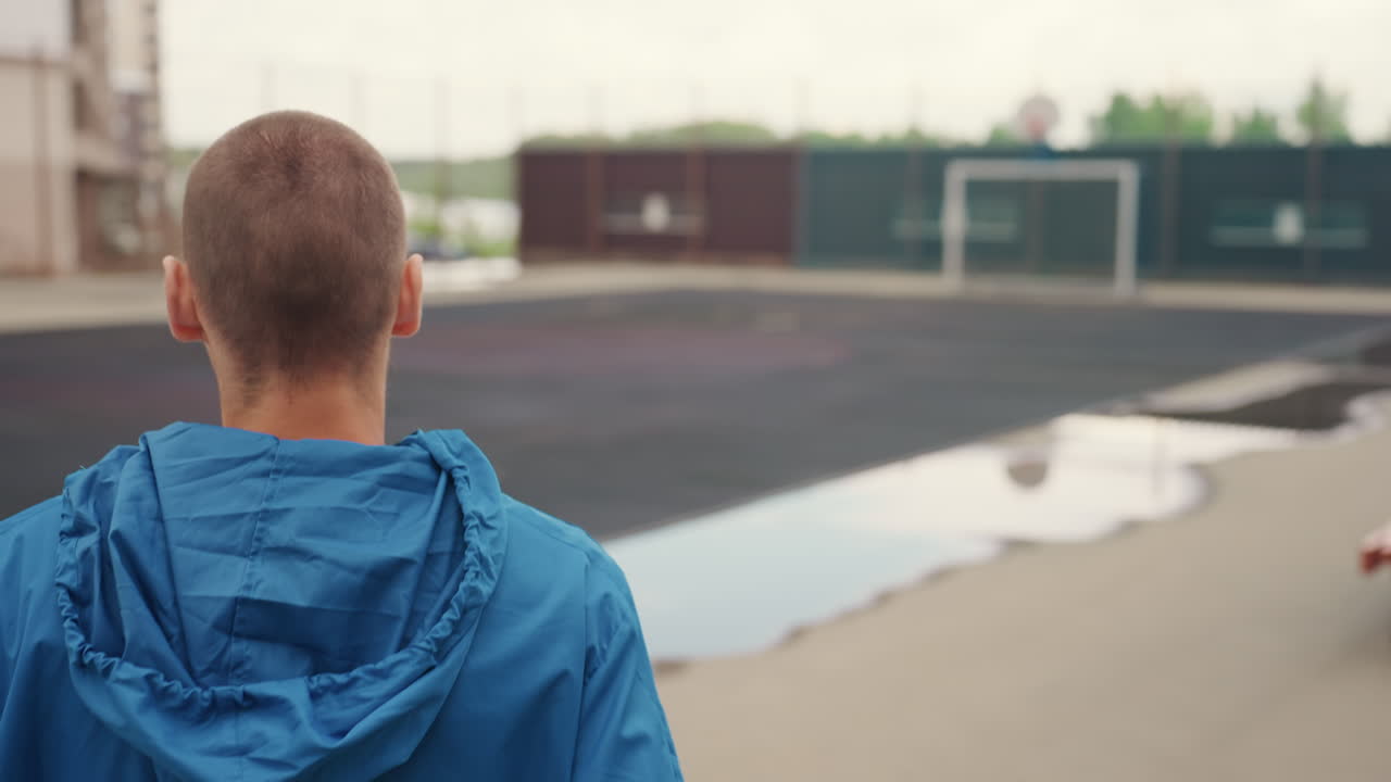 Blanke jongen met blauw jack bij hek; eenzame jonge blanke atleet pauzeert voor een afgesloten stedelijk voetbalveld, inspecteert het natte veld en de plassen na de regen, capuchon op en handen in zijn zakken, reflecterende houding