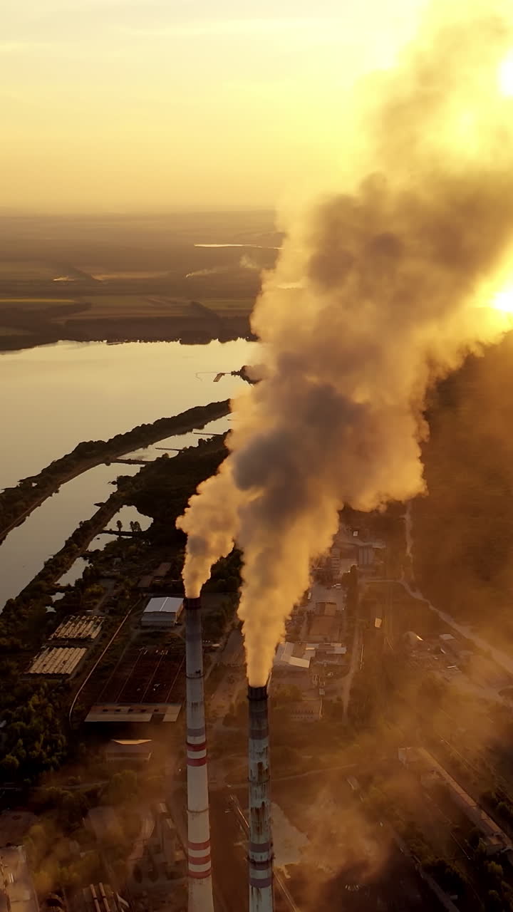 Power factory in nature at sunset. Thick smoke coming out from industrial pipes on the evening river background. Polluting the atmosphere. Aerial view. Vertical video