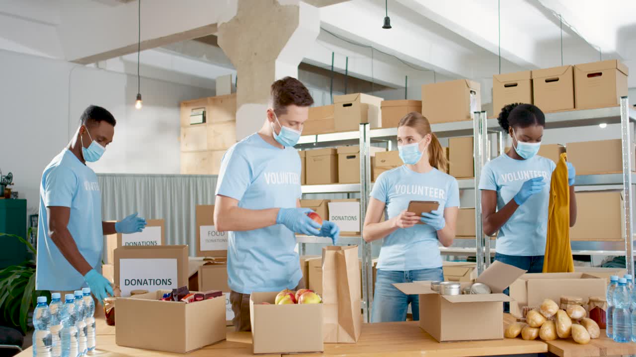 многонациональная группа волонтеров в масках для лица упаковывает коробки с едой и одеждой на благотворительном складе