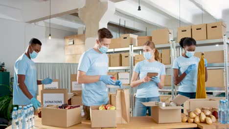 multiethnic group of volunteers in facial mask packing boxes with food and clothes in charity warehouse