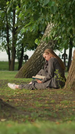 lady in grey clothing sits outdoors under tree reading book with hand on page, slightly bent forward, leg stretched, the background features greenery and people walking in the distance