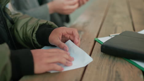close up of hands folding paper into kite shape on wooden table, showing detail of crafting process as two people work together in creative activity outdoors with focus on teamwork and learning