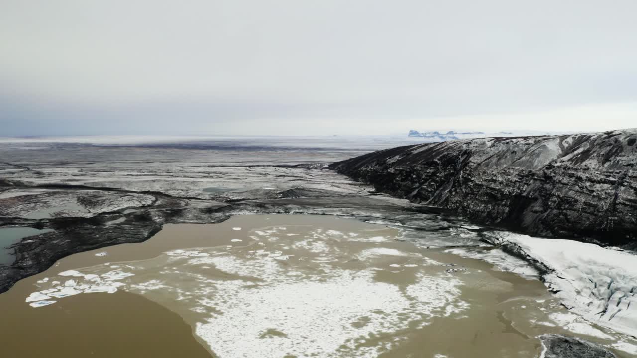 Aerial shot highlighting rugged cliffs, icy patches, and a muddy lagoon in the remote landscape of Svinafellsjokull.