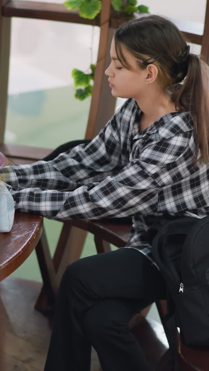 십 대 소녀가 쇼핑몰 아트리움의 나무 테이블에 앉아 가방에서 물건을 찾고 있어요. 빨간색 휴대폰 케이스와 반짝이는 파우치를 가방에서 꺼내고 있으며, 세심한 표정으로 체크무늬 셔츠를 입고 포니테일을 하고 있어요. 늘어진 녹색 식물이 보이고 자연스러운 모습입니다.