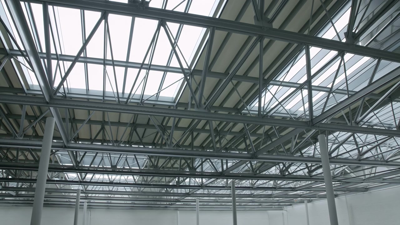 Low angle view inside empty industrial hall with exposed steel trusses, rectangular skylights, repeating beams, and vertical support columns, creating strong geometric structure, cool daylight, and