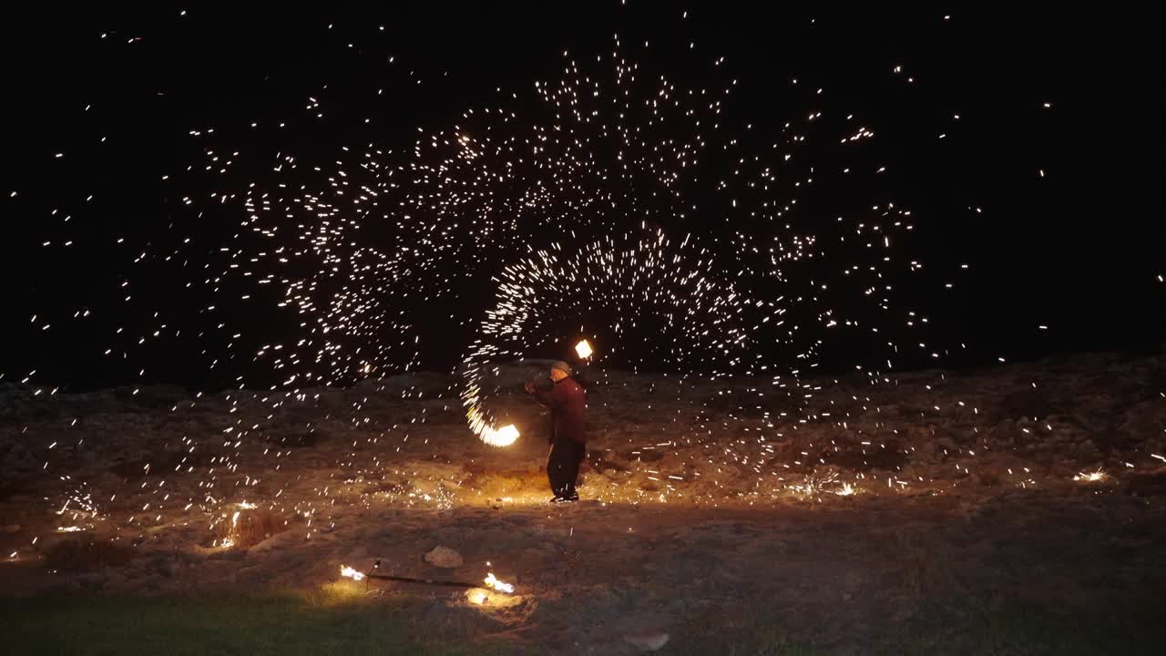Un artista fa roteare dei bastoni infuocati, creando scintille luminose durante un evento all'aperto nel parco di notte. Il pubblico osserva con meraviglia mentre le fiamme illuminano il cielo buio