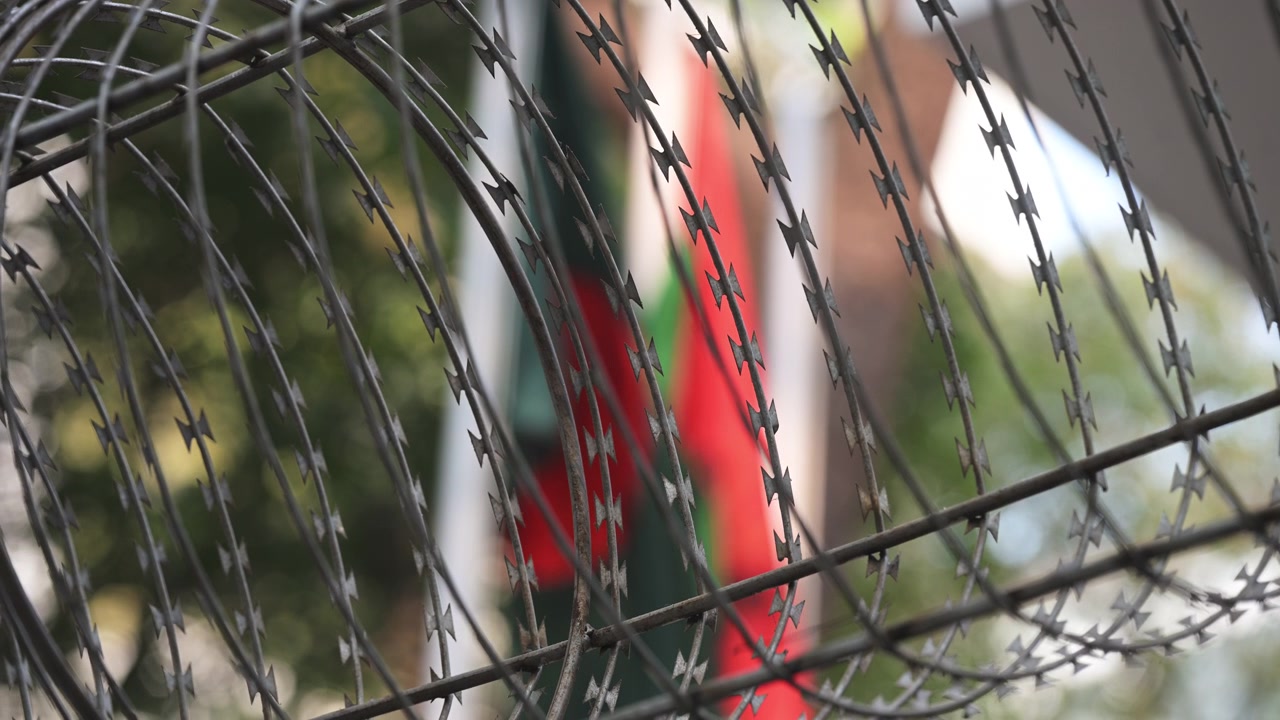 Coiled security razor wire stands in sharp focus as the BNP party flag hangs obscured in the soft background in Dhaka. Documents regional political conflicts, party crackdowns, and safety.