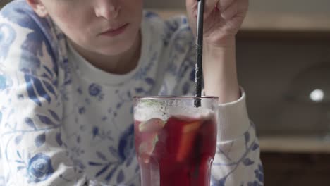 young girl is stirring a red lemonade in a glass with plastic straw, pushing down mint leaf