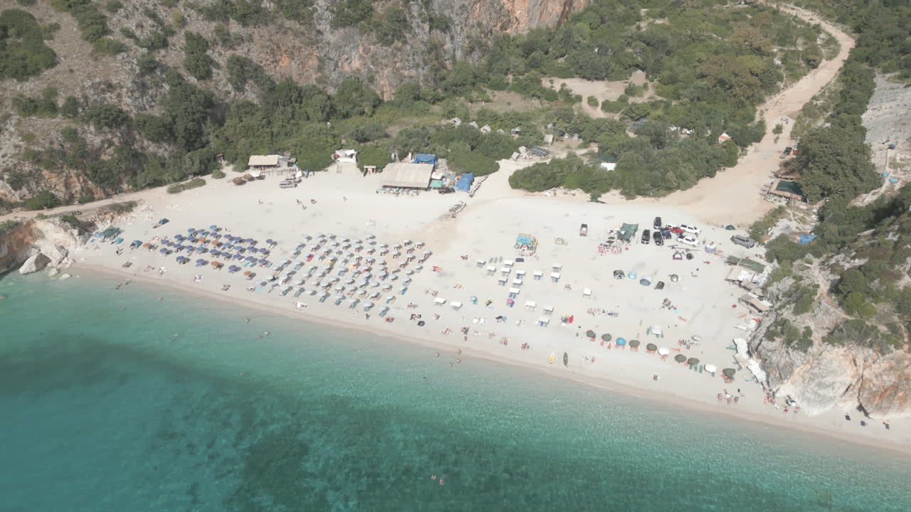 Drone shot over Gjipe beach Albania with tourists on the beach and clear blue water near the mountains and the sea on a sunny and bright day and green nature around with sun beds towels LOG