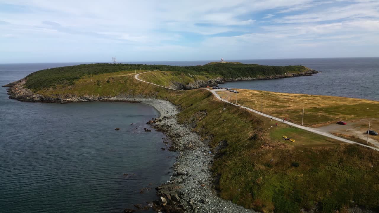 A drone soars over Ferryland, showcasing a narrow causeway linking the mainland to a rocky peninsula, with blue waters on both sides and grassy cliffs