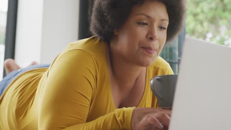 video of happy plus size african american woman lying on floor with coffee and using laptop
