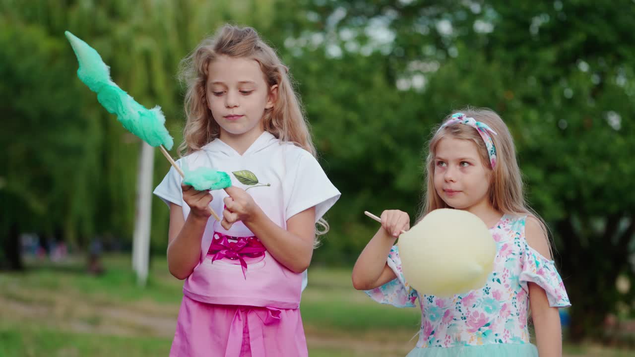 Two little girls eating cotton candies outdoors. Happy kids enjoys eating colorful sweet candy-floss in the summer park. Happy childhood.