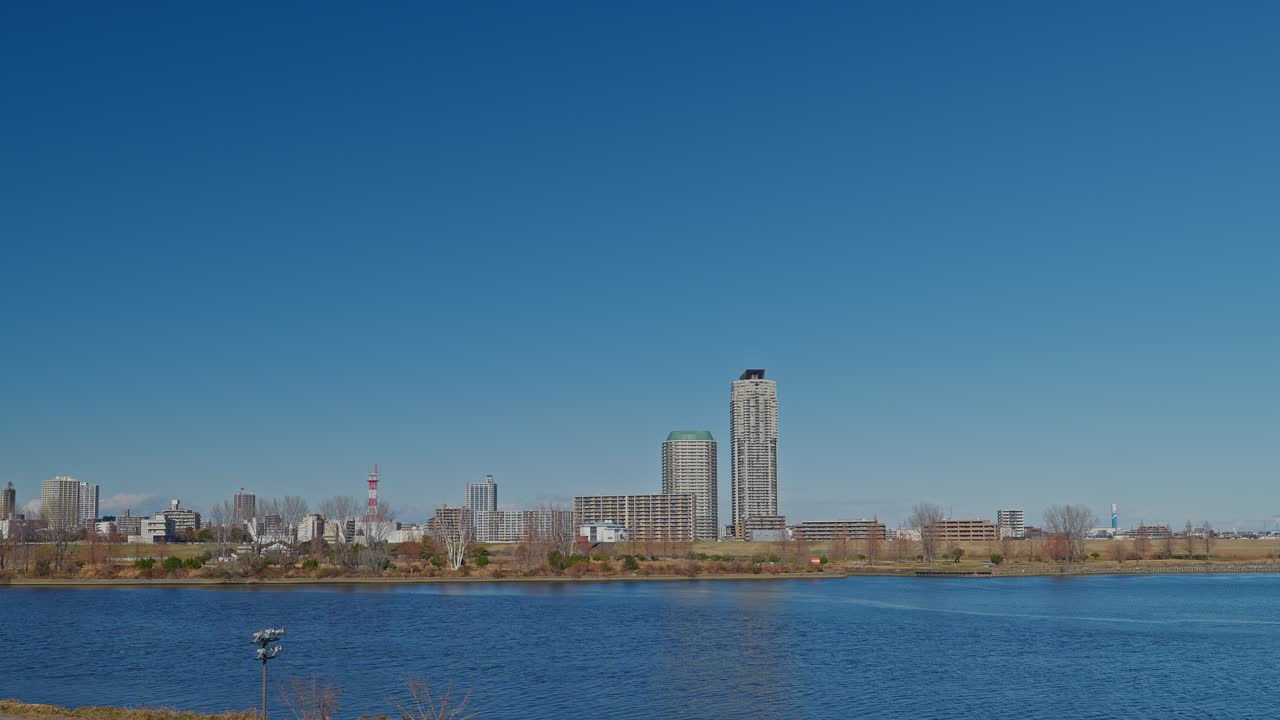 Uma ampla vista panorâmica do Rio Arakawa no Japão, olhando em direção aos arranha-céus residenciais da Cidade de Kawaguchi. O rio azul está sob um céu vasto e claro com margens relvadas abertas.