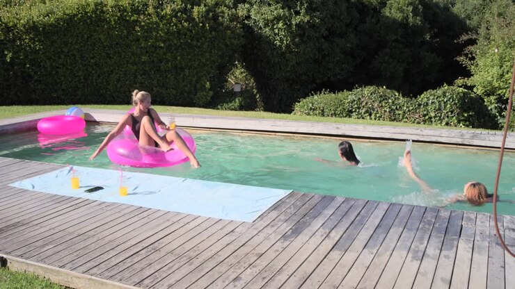 Female friends racing across lawn in swimwear jumping into pool swimming to pink float with drink
