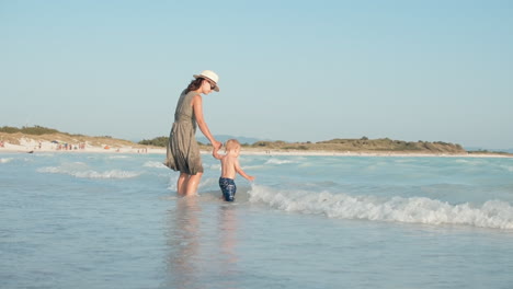 active son leading mother to waves at seaside. woman and boy standing in water.