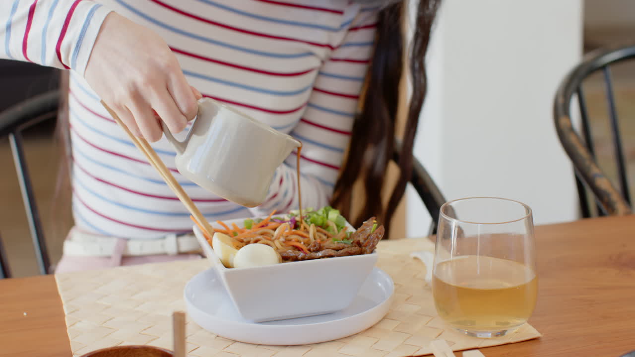 Pouring sauce over noodles, woman enjoying meal with drink at dining table