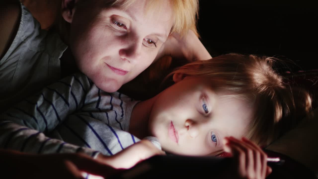엄마와 딸이 함께 침대에 누워 어둠 속에서 태블릿 화면을 보고 있다
