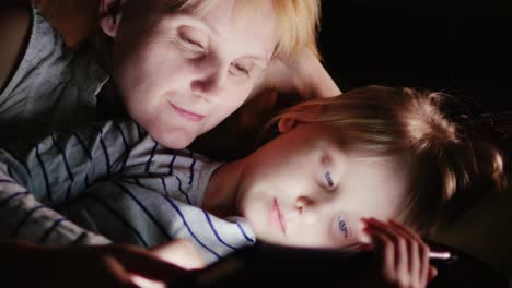 Mutter-Und-Tochter-Liegen-Zusammen-Auf-Dem-Bett-Und-Schauen-Im-Dunkeln-Auf-Den-Bildschirm-Des-Tablets