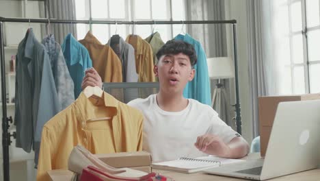 asian man online seller recommending a shirt while using computer for selling clothes at home