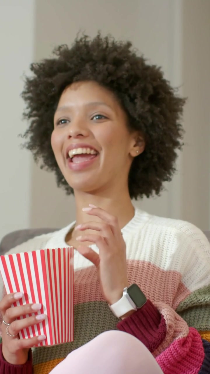 Video verticale: Donna in maglione a righe che si allunga per prendere una vaschetta di popcorn sul divano, reagendo a uno spettacolo.